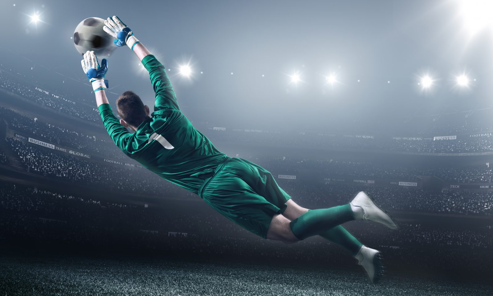 Soccer goalie jumping in motion for a ball while defending his gates. The stadium is blurred behind him. Only the lights of the stadium shine brightly, creating a halo effect around the bulbs. A player is wearing unbranded soccer uniform.