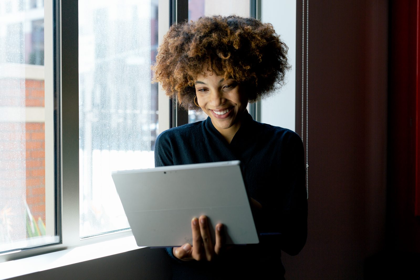 Female college student standing inside building near windows, smiling and using Surface Pro (screen not shown).