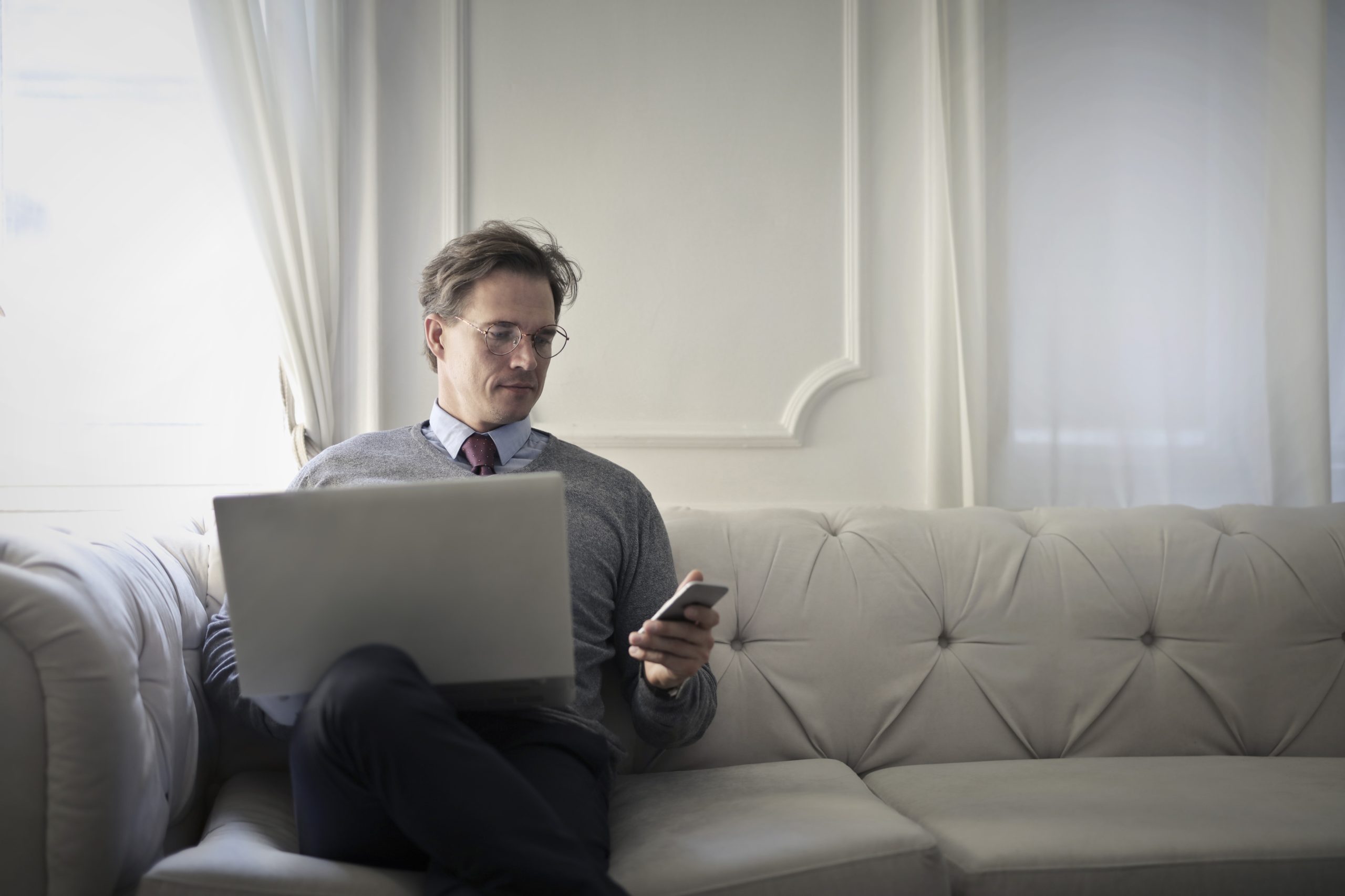 A man in a shirt, tie and jumper sits on a sofa at home with a laptop on his knees, and holding a mobile phone