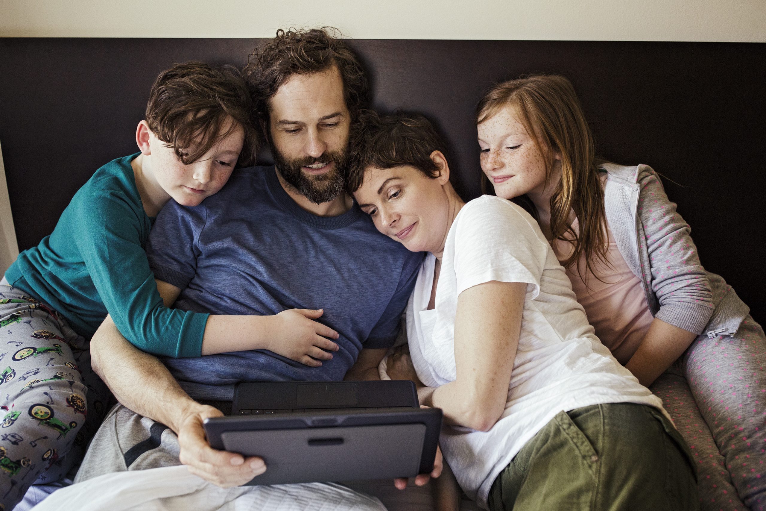 A family with mum, dad and two children look at a tablet while sitting on the sofa.