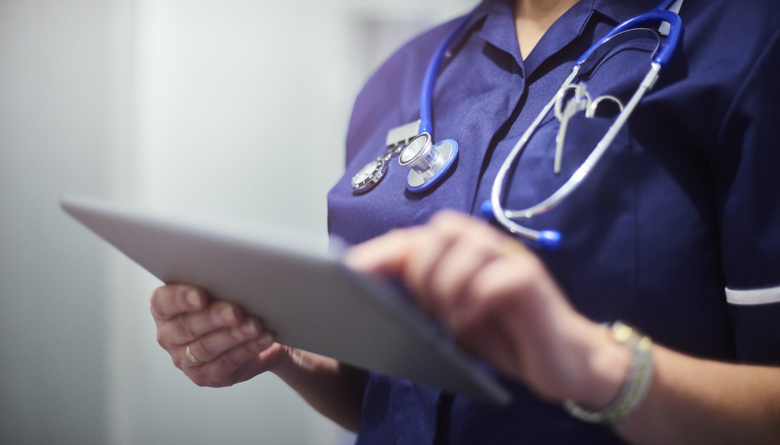 Female nurse typing on digital tablet in hospital or surgery. She is wearing a dark blue nurse’s top and has her stethoscope around her neck. She is looking at her patients records on her digital tablet.