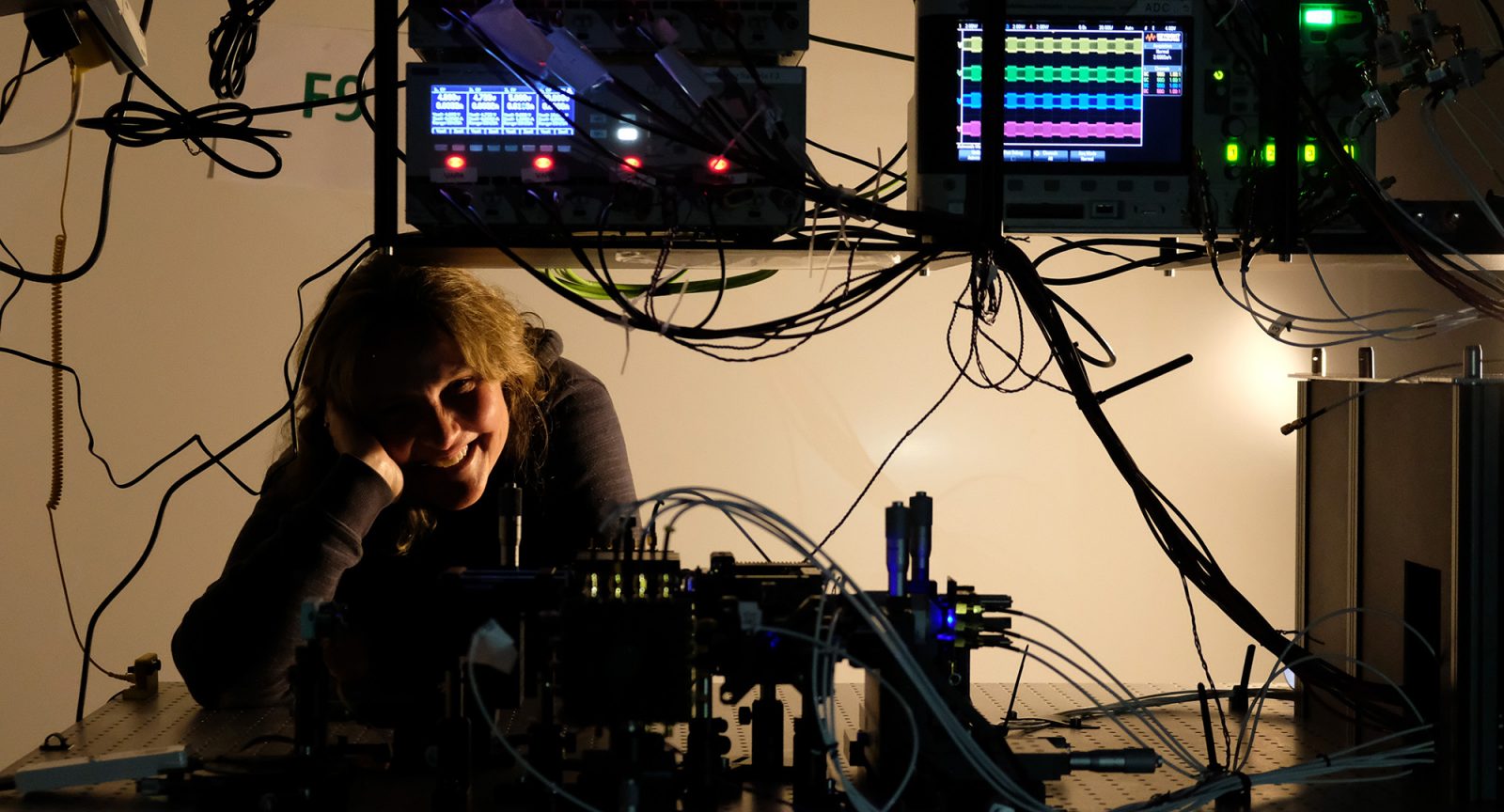A woman is looking out underneath some computers with a lot of wires and lights. She is smiling with her head in her hand.