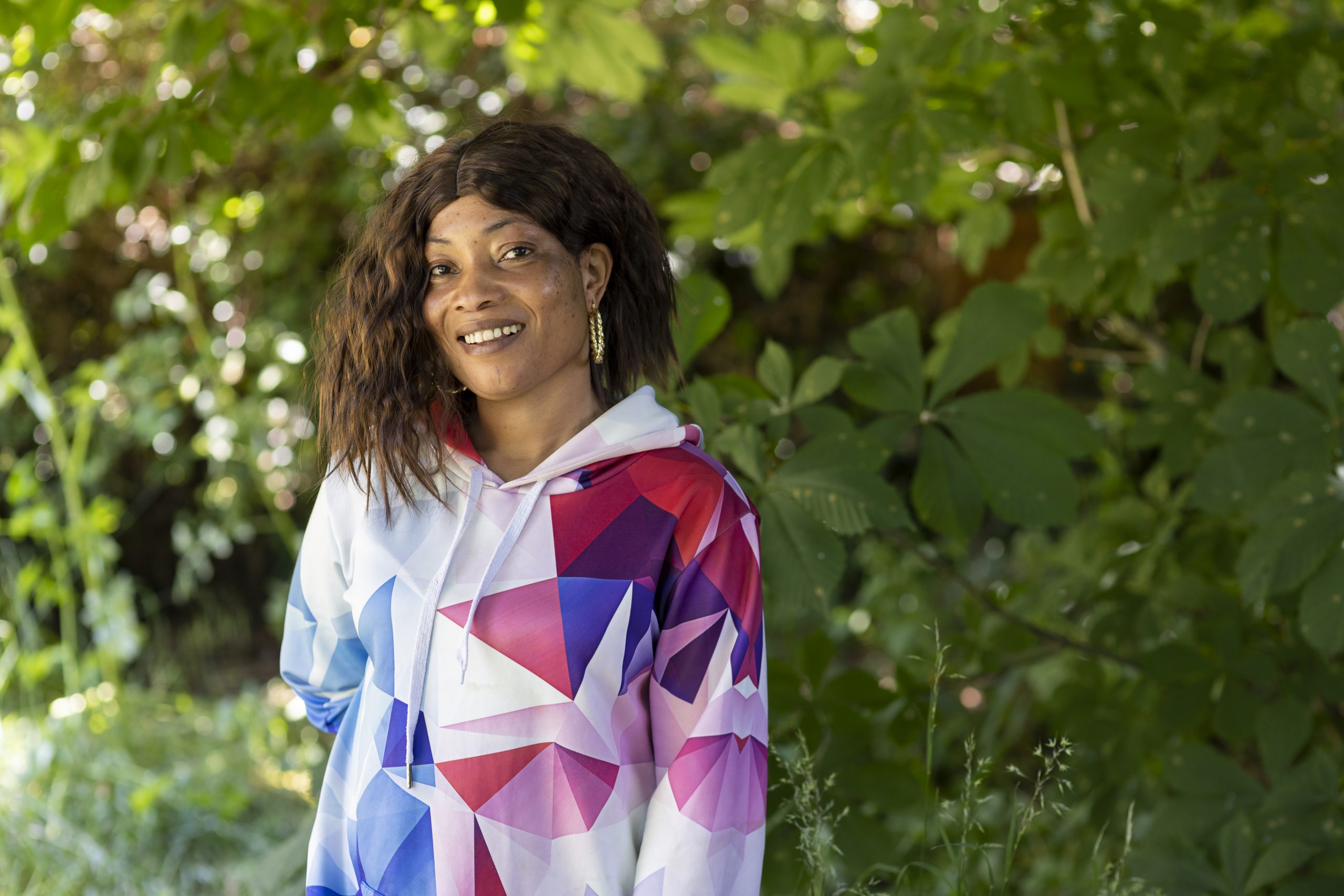 A smiling woman in a white hoodie with colourful geometric patterns in purple, pink and blue, standing in front of a tree.