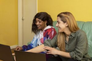 Two women sit on a sofa and look at two laptops during a lesson on digital skills