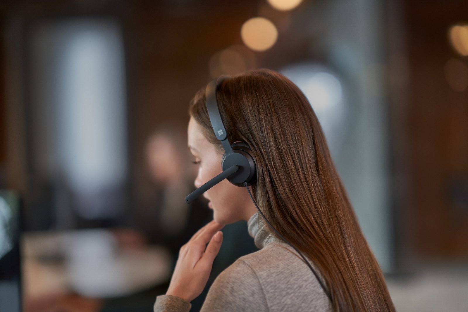 A woman with long dark hair wears a headset. She has her back to the camera and is looking at a computer screen.