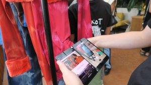 A photo of a woman using a phone (a Microsoft Surface Duo) to take a photo of a QR code on a label of a pink and orange Ahluwalia dress.