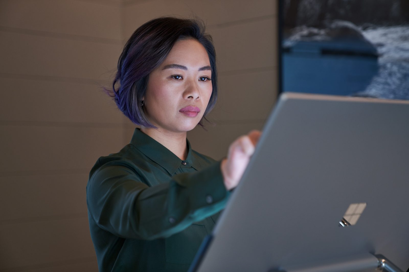 Side profile of a woman wearing a dark shirt in a dim office reaching up and working on a Microsoft Surface Studio. Keywords: touch screen, desktop, cloud security, threat protection, secure score, monitoring, Microsoft Security collection