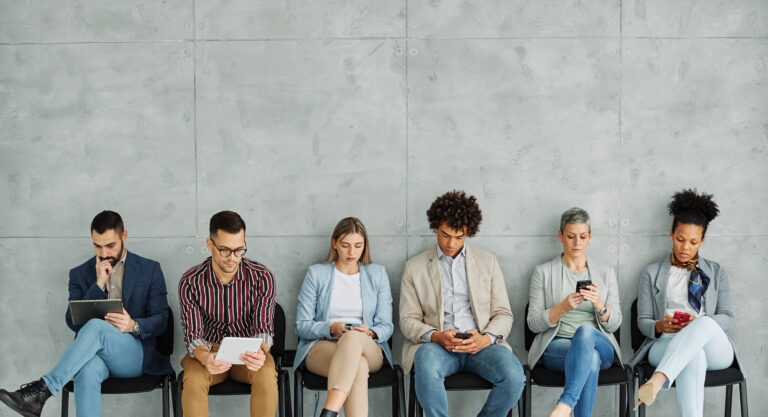 Group of young job applicants sitting waiting for an interview and looking at their smartphones and tablets