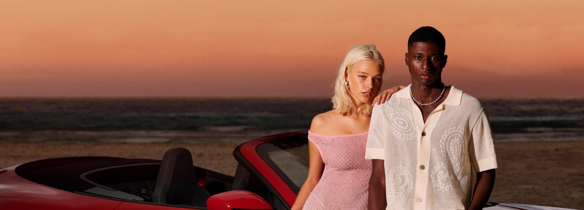 Young woman and man standing next to red sports car on beach at sunset