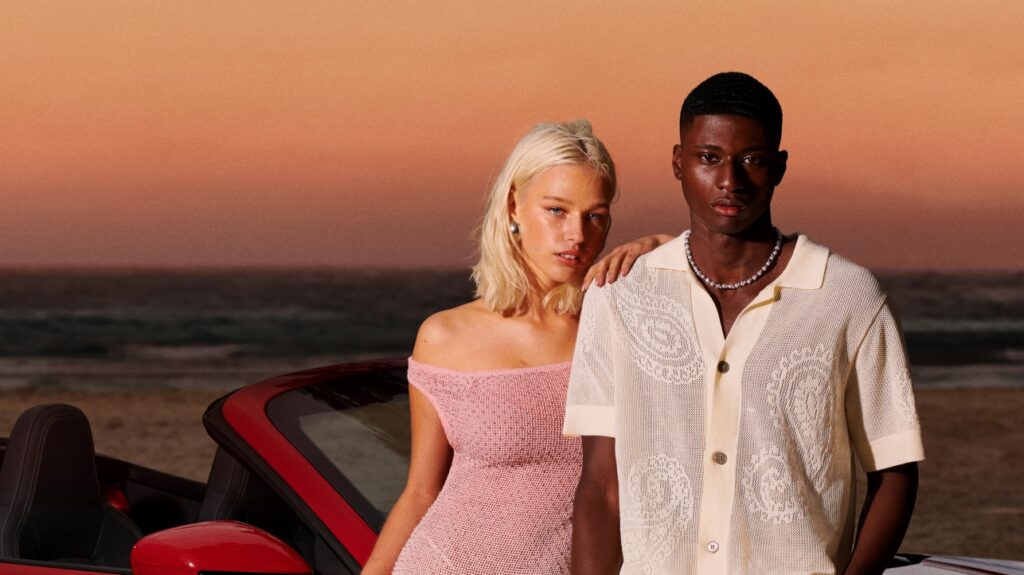 Young woman and man standing next to red sports car on beach at sunset