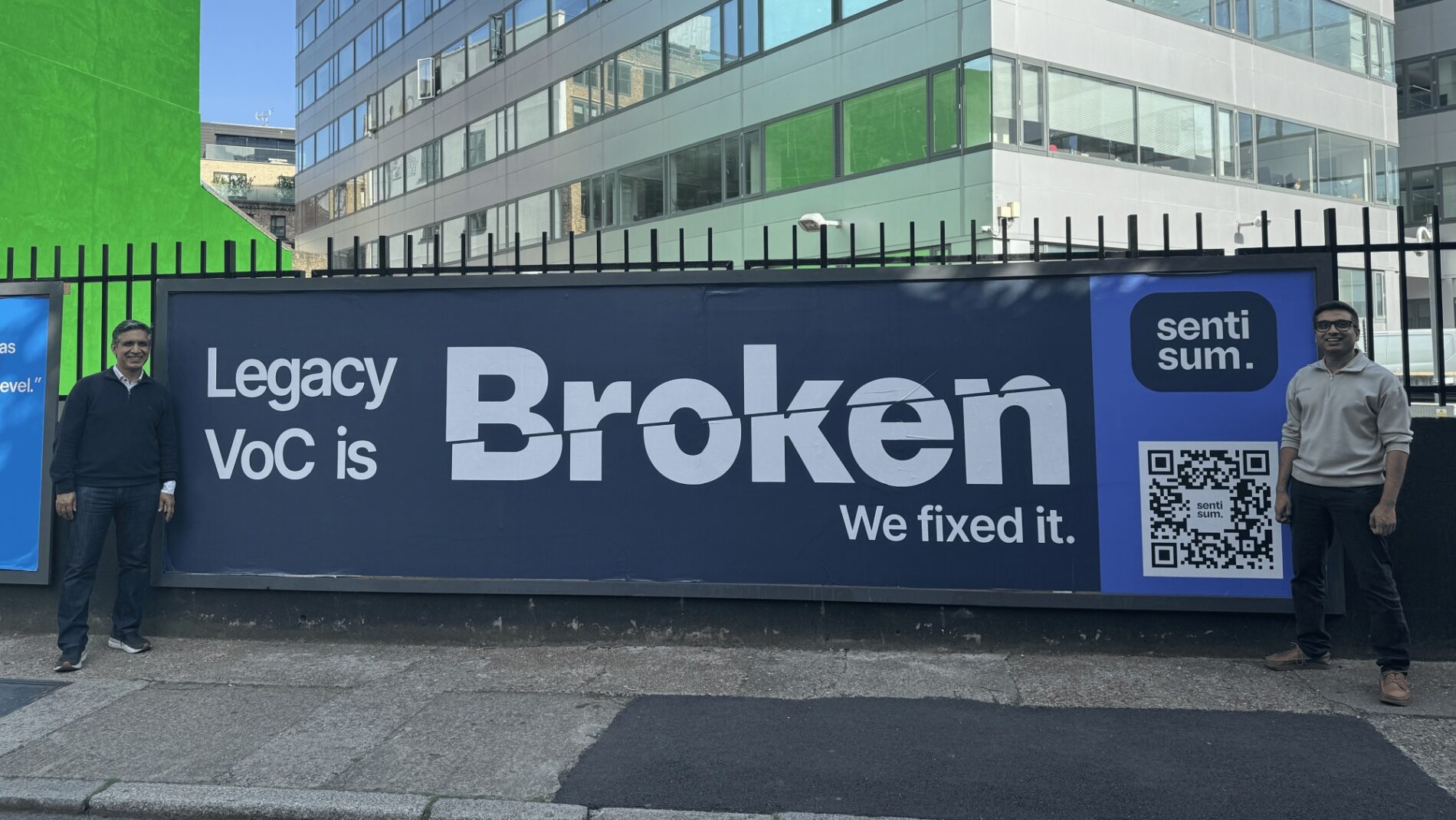 SentiSum founders Vivek Ganotra (left) and Sharad Khandelwal standing in front of a billboard saying: 'Legacy VoC is Broken. We fixed it.'