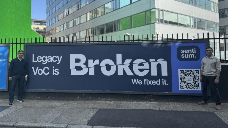 SentiSum founders Vivek Ganotra (left) and Sharad Khandelwal standing in front of a billboard saying: 'Legacy VoC is Broken. We fixed it.'