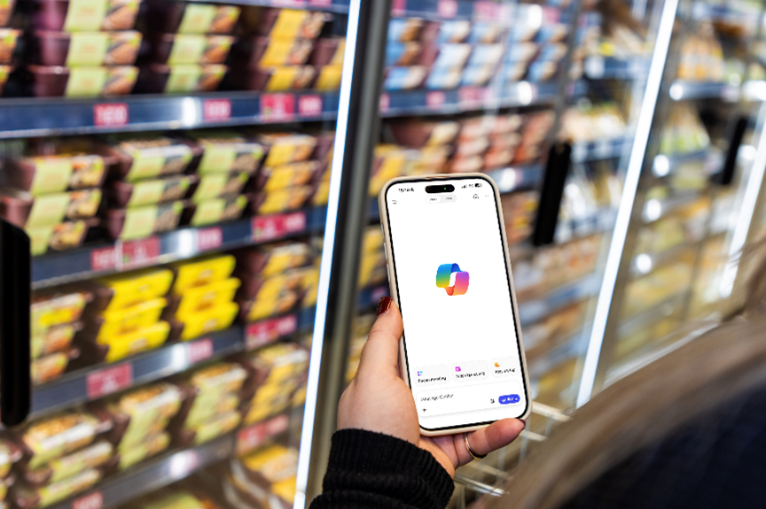 Close-up of hand holding a smartphone showing the Copilot logo on screen with chiller cabinet in the background