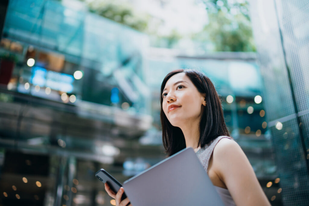 Successful and modern young Asian businesswoman carrying smartphone and laptop symbolising human face of recruitment